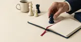 A hand setting a navy chess knight on an open notebook with a red ribbon