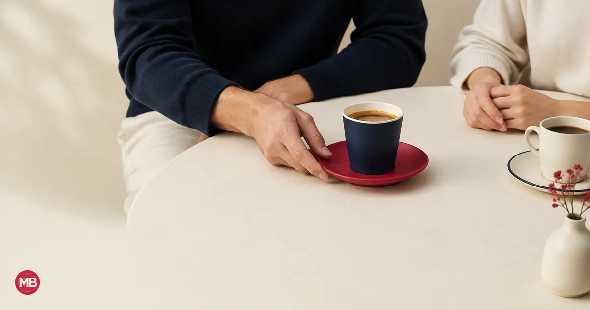 Two people at a café table, one sliding a navy cup on a red saucer to the other